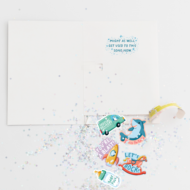 Opened Joker Greeting endless musical baby shower card showing large glitter and colorful baby-themed stickers scattered around with special delivery truck, shark, rocket and other fun designs