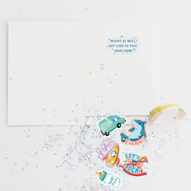 Opened Joker Greeting endless musical baby shower card showing large glitter and colorful baby-themed stickers scattered around with special delivery truck, shark, rocket and other fun designs