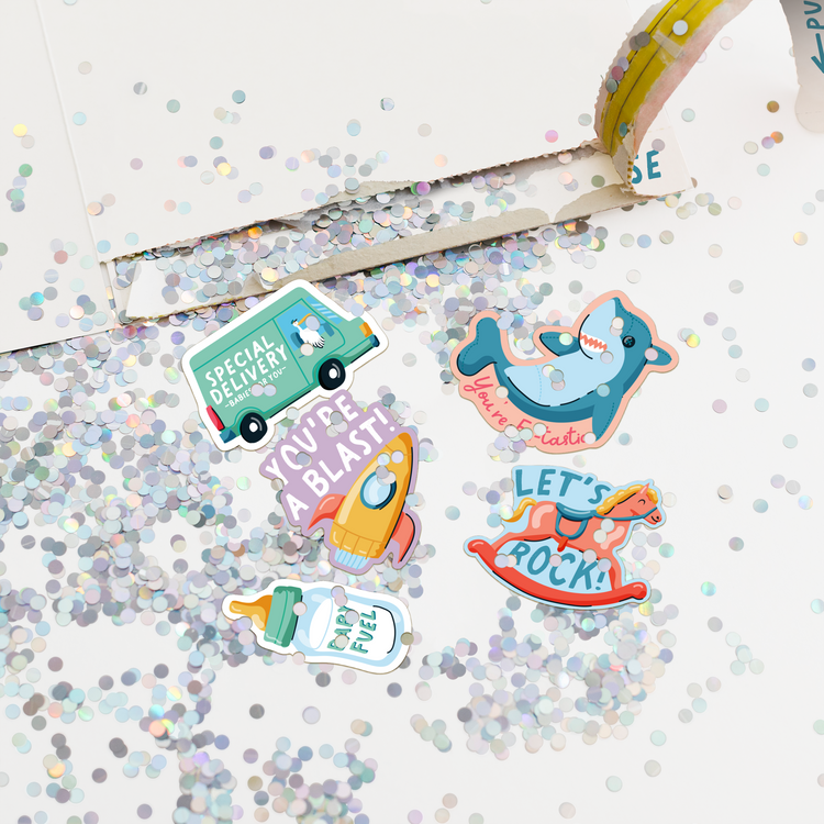 Close-up of opened Joker Greeting baby shower card showing large glitter explosion and colorful baby stickers including special delivery truck shark lets rock rocking horse and baby bottle