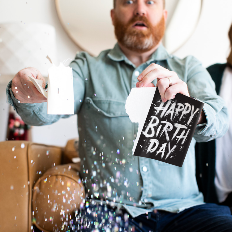 Glitter Action Shot:
Man holding Birthday Screamer musical prank card with chunky glitter spilling everywhere showing black design with white paint splatter typography and hidden glitter surprise feature