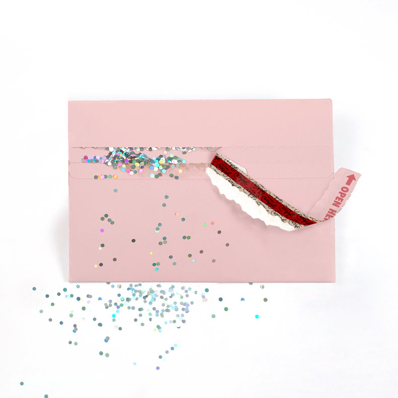 (Envelope being opened with glitter spilling action): Blush pink glitter trap envelope mid-opening with Joker Greeting branding visible and multicolored glitter creating romantic prank chaos
