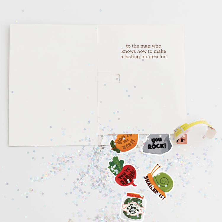 Open Father’s Day greeting card with message “to the man who knows how to make a lasting impression,” surrounded by scattered glitter and colorful pun stickers revealed from a hidden pull tab labeled “pull my finger.”