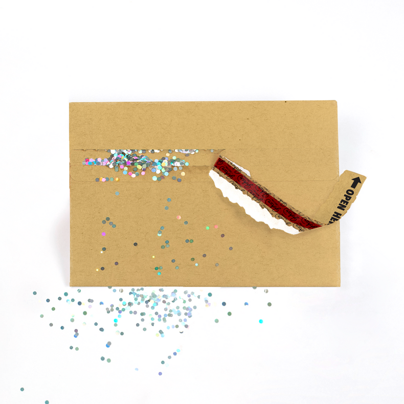 A brown kraft envelope with a strip of glitter on the side that has been pulled to dispense multicolored glitter onto the surface.