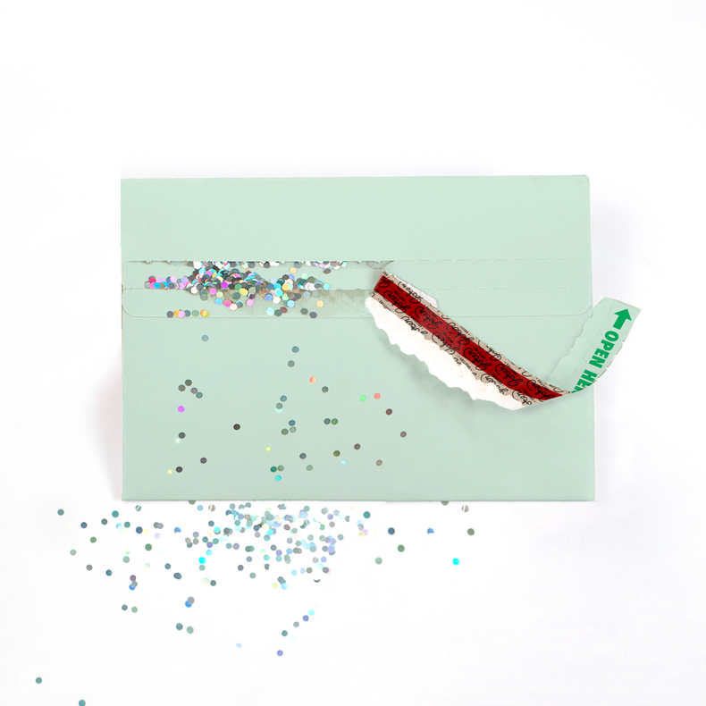 (Envelope being opened with glitter spilling): Mint green glitter trap envelope being opened with Joker Greeting branding visible and colorful glitter spilling out creating surprise prank