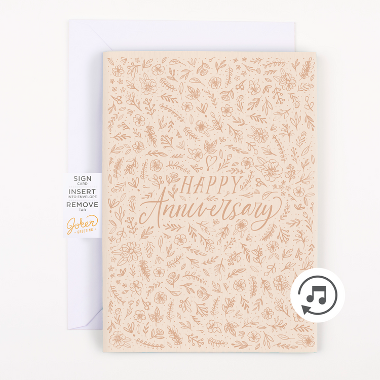 A greeting card with a floral pattern and the text 'Happy Anniversary' in a cursive font, with a small piece of glitter visible at the bottom right corner, presented alongside a plain white envelope.