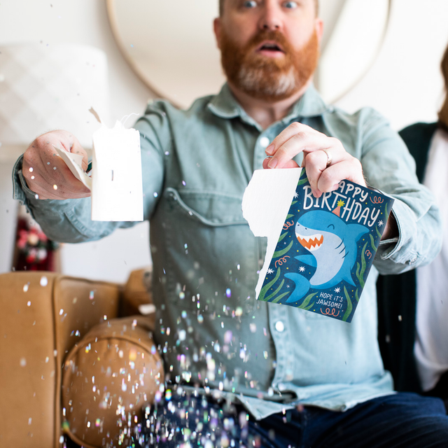 Glitter Action Shot:
Man holding Baby Shark musical birthday prank card with colorful glitter spilling everywhere demonstrating glitter release when card is broken