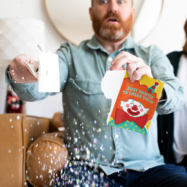 Man holding opened Joker Greeting endless musical cheer up card with large glitter explosion effect and surprised facial expression