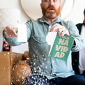 Man holding torn Feliz Navidad card with glitter explosion showing endless musical prank activation in living room setting