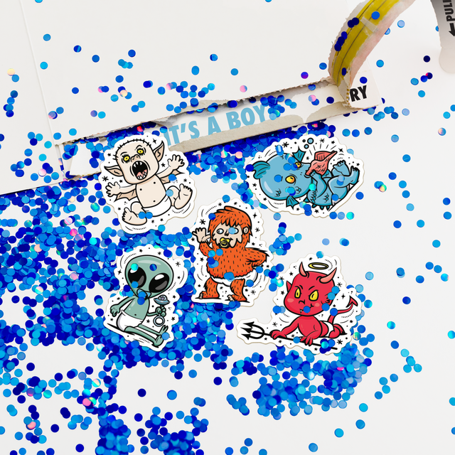 Open gender reveal card revealing IT'S A BOY message with blue glitter and character stickers including alien, monster, and demon designs