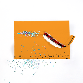 (Envelope being opened with glitter spilling): Orange Halloween glitter trap envelope being opened with colorful glitter spilling out creating spooky prank surprise