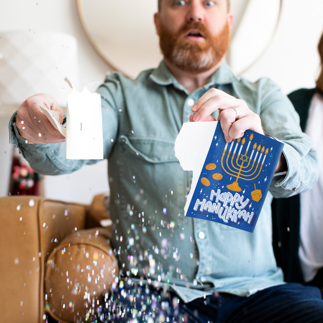 Person holding activated Joker Greeting Hanukkah card with Happy Hanukkah menorah design and large glitter falling showing endless musical prank in action