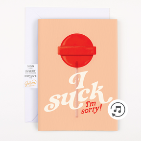 A greeting card with a graphic design showing a red lollipop with the text 'I suck, I'm sorry!' on an orange background. A plain white envelope is included.