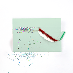 (Envelope being opened with glitter spilling): Mint green glitter trap envelope being opened with Joker Greeting branding visible and colorful glitter spilling out creating surprise prank