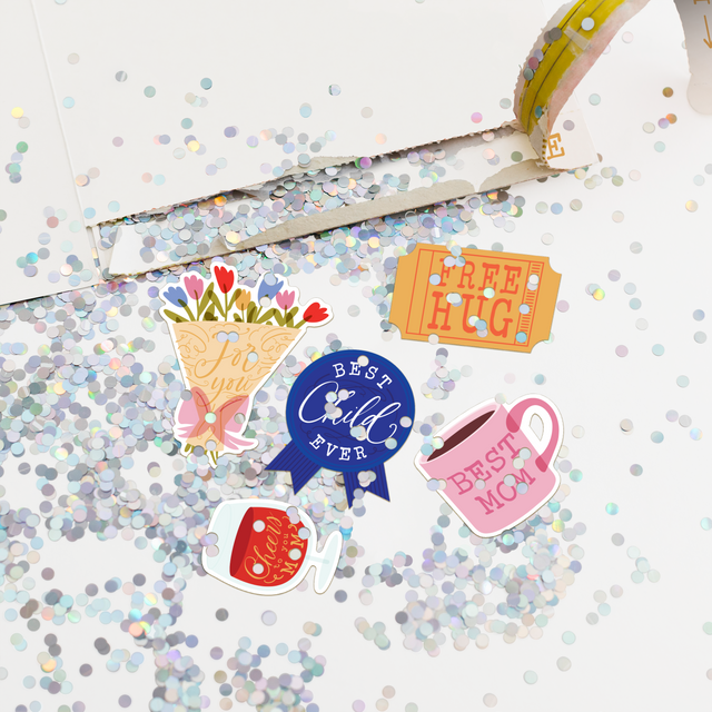  Torn open Mother's Day card revealing large glitter release and Mother's Day themed stickers including flower bouquet Best Mom mug and Best Child Ever ribbon scattered everywhere