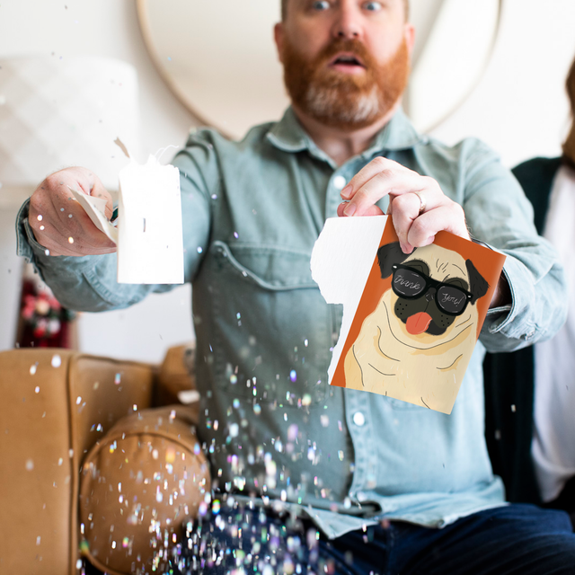 Person holding torn pug musical greeting card with surprised expression as large glitter falls around them demonstrating glitter activation
