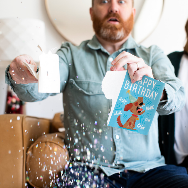 Glitter Action Shot:
Man holding Yappy Birthday dog musical prank card with chunky glitter spilling everywhere showing cartoon dog illustration and barking sound effects design